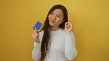 Sarı arka planda kredi kartı tutan gülümseyen bir kadın pozitifliğini ve güvenini dile getiriyor.