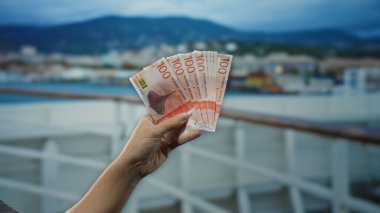 Arka planda dağlarla dolu bir gemi güvertesinde Norveç kronunu tutan adam bulutlu bir gökyüzünün altında açık hava seyahat teması sergiliyor..