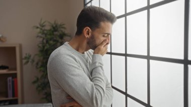 Oturma odasındaki bir pencerenin yanında duran genç adam, düşünceli ve endişeli görünüyor, içinden güneş ışığı akıyor, evin barışçıl ama düşünceli atmosferini arttırıyor..