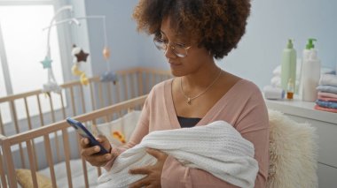 Kıvırcık saçlı bir kadın, yatak odası ortamında akıllı telefon kullanırken bebeği kucaklıyor. Modern annelik ve kapalı mekan atmosferinin bir karışımını sergiliyor..