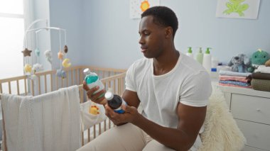 Genç adam yatak odasında oturup bebek beşiğinin yakınındaki şişeleri inceliyor. Ev içinde rahat bir yerde, dikkatli ebeveynlik ve modern yaşam tarzını ön plana çıkarıyor..