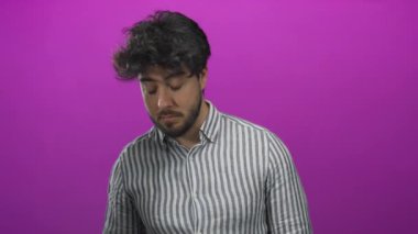 Sakallı ve çizgili gömlekli genç adam canlı pembe arka plana karşı dikkatle dinliyor, merakını iletiyor ve modern bir ortama odaklanıyor..