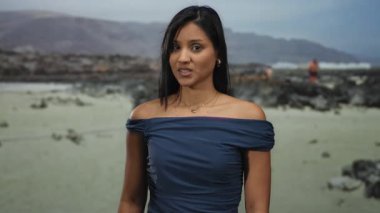 Genç Latin kadın deniz kenarında aptalca bir jest yapıyor. Sahilde neşeli bir ruh hali sergiliyor. Arka planda dağlar, açık mavi gökyüzünün altında..