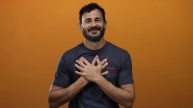 Sakallı İspanyol sakallı adam canlı turuncu bir arkaplan karşısında içten bir şekilde gülümsüyor. Elleri kalbimde. Minnettarlığı ve pozitifliği simgeliyor.