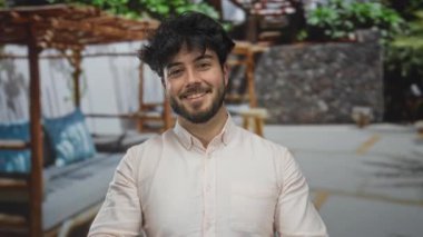 Güneşli bir günde bardakları ayarlayan genç İspanyol bir adam, arka planda yeşil yapraklı günlük gömleğiyle gülümsüyor, pozitif ve neşeli bir ruh hali içinde..