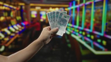 Arka planda renkli kumar makineleri olan canlı bir kumarhane ortamında paralar tutan adam..