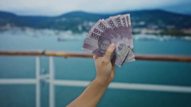 El ele tutuşan Endonezya rupiah banknotları, deniz kenarı arka planı, tekneler ve okyanus manzaralı bir limanın yakınında para sergileyen adam, seyahat ve finans temalarını öneriyor..