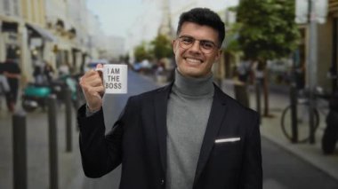 Genç adam elinde bir kupa tutarken gülümsüyor 'Ben patronun metni, canlı bir şehir caddesinde duruyor arka planı bulanık, kendine güveni ve tarzı açık havada.