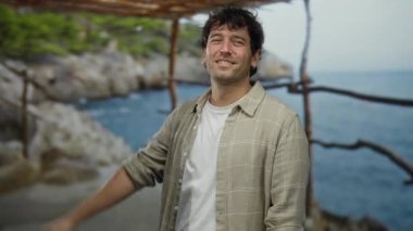 Denizin kenarında dikilen adam gülümsüyor, günlük giysiler giyiyor, güneşli bir gökyüzünün altında arka planda kayalar ve yeşilliklerle plaj manzarasının tadını çıkarıyor..