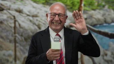 Takım elbiseli son sınıf öğrencisi elinde mikrofonla sahil kenarında el sallıyor ve gülümsüyor. İş dünyası ve medya, çok zarif bir jestle ortam sunuyor..