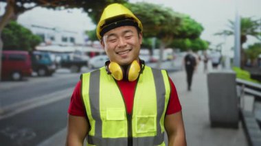 Güvenlik teçhizatı ve sert şapkalı güler yüzlü adam şehir yaşamının ortasında inşaat sektöründe profesyonelliğini sergileyen kalabalık bir caddede kendinden emin duruyor..
