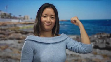 Genç bir kadın, deniz kenarında kaslarını kasıyor. Mavi okyanus dalgalarıyla sahilin manzarasına karşı güç ve güç gösterisi yapıyor..