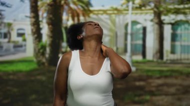 Parkta boynu ovalayan bir kadın ağaçların yanında acı çekiyor ve parlak güneş ışığında kemerler takıyor beyaz tanklar giyiyor orta boy doğal saç pozu veriyor.