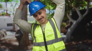 Yansıtıcı yeleği ve kasketi olan genç bir adam şehir dışındaki bir sokakta elleri ile kalp hareketi yaparak şehir çalışma ortamında neşelendirici bir an yakalıyor..