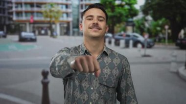 Şablon gömlekli genç İspanyol adam şehrin arka planında güneşli bir gökyüzünün altında yumruğuyla güvenle gülümsüyor..