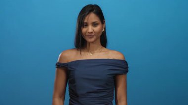 Genç bir kadın, seni izlediğini işaret ediyor, kendine güvenerek gülümsüyor, mavi bir duvarın arkasında, koyu renk bir elbise giyiyor, oyuncu ve gözlemci bir ifade sergiliyor..