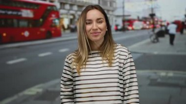 Kırmızı otobüslerle dolup taşan bir caddede sarı saçlı genç bir kadın, ikonik şehir atmosferi ve günlük moda ile canlı bir şehir manzarası sergiliyor..