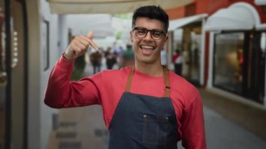 Canlı bir sokak ortamında kırmızı bir kazak ve kot önlük giymiş, kendine güveni ve modern şehir tarzı açık hava tarzıyla gülümseyen genç İspanyol bir adam..