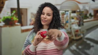 Dışarıda elinde kırmızı bir kalple gülümseyen ve şehir caddesinde renkli süveter giyen bir kadın..