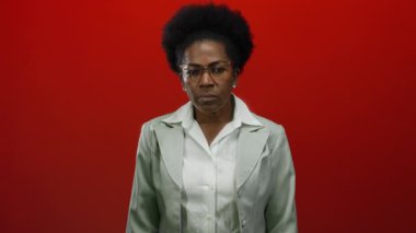 Gözlüklü bir kadın, canlı kırmızı bir arka planda, beyaz bir bluz ve takım elbise giyiyor, düşünceli bir şekilde düşünüyor ve kendine güveniyor..