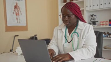 Klinikte çalışan bir kadın, steteskop ve sağlık çizelgesiyle tıbbi malzemeler ve anatomi posterleriyle çevrili profesyonel sağlık hizmetleri ortamı sergiliyor..