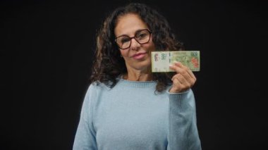 Arjantinli banknotları siyah arka planda tutan orta yaşlı bir kadın stüdyo benzeri bir ortamda göze çarpan bir para birimi gösteriyor..