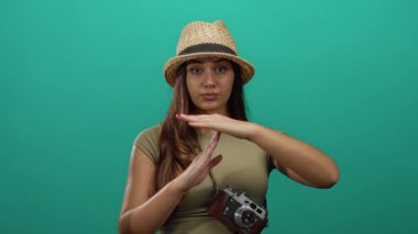 Yeşil arka planda poz veren şapkalı bir kadın boynunda bir kamerayla izleyiciye güvenle bakarken el kol hareketi yapıyor.