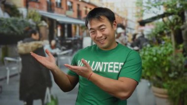 Kentsel terasta gülümseyen Asyalı bir adam, yeşil tişörtlü, çevresi açık kafe dekorları ve yeşilliklerle çevrili, dostane ve misafirperver bir atmosfer öneriyor..