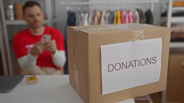 Yardım odasında bağış kutusuyla telefon eden, gönüllü tişörtü giyen, elbiselerle çevrili, cömertliği ve sosyal yardımı sembolize eden genç bir adam..