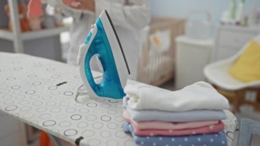 Çamaşırlarını yıkama odasında, katlanmış giysiler ve temiz ve düzenli bir mekanla çevrili mavi buhar ütüsüyle dolu ütü masasının yanında sıralayan bir kadın..