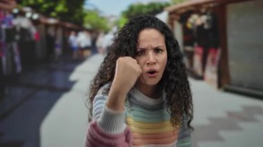 Kıvırcık saçlı bir kadın hareketli bir şekilde açık hava pazarında, yoğun müşterilerin arasında renkli bir süveter giyerken güçlü duygular taşıyor..