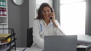 Hastane ofisinde steteskopla telefonla konuşan kadın doktor dizüstü bilgisayardaki hasta kayıtlarıyla konuşurken gülümsüyor..