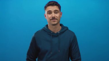 Kapüşonlu genç İspanyol adam kollarını izole edilmiş mavi duvara yaslıyor. Rahat ve rahat davranışlar sergiliyor.