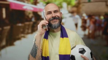 Futbol topu tutan ve şehir merkezinde telefonla konuşan yakışıklı kel adam sarı mor atkı takıyor spor tutkusu ve bağlantısını gösteriyor..