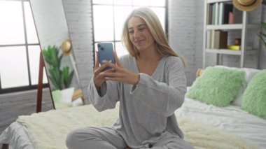 Kadın yatakta oturmuş video çekiyor, gülümsüyor ve yatak odasında el sallıyor. Modern dekor ve güneş ışığı geniş pencerelerden içeri yayılıyor..