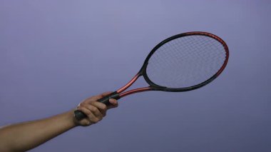Mor izole edilmiş arka planda tenis raketi tutan erkek el, spor ve eğlence unsurlarını vurguluyor.