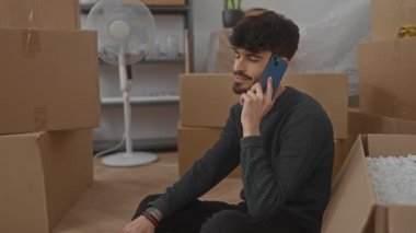 Genç adam oturma odasında karton kutularla konuşuyor. Yeni eve taşınırken, yerde rahatça oturuyor..