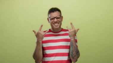 Gözlüklü genç İspanyol adam, sarı arka planda, dövmeli kollu ve çizgili gömlekli taş jestler yapıyor. Ayrı bir kutlama sırasında enerjik ve neşeli bir hava yayıyor.