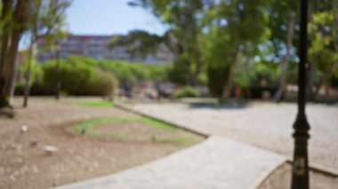 Bulanık görüntü, güneşli bir günde çekilmiş ağaçlı, patikalı ve arka planda bir bina olan bir açık hava parkını gösteriyor..