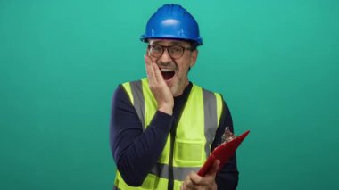 Güvenlik teçhizatı içindeki olgun bir adam, parlak yeşil bir arka plana karşı komik bir şekilde tepki veriyor. Elinde bir pano var, gözlük ve şapka takıyor..
