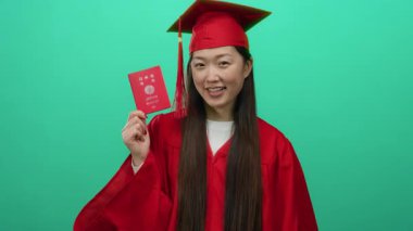 Kırmızı mezuniyet elbisesi içindeki Japon kadın yeşil arka planda Japon pasaportunu gururla sergiliyor, eğitim başarısını ve uluslararası seyahat fırsatlarını simgeliyor..