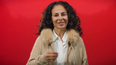 Orta yaşlı, kıvırcık saçlı, sırıtan, izole edilmiş kırmızı bir arka plana başparmak sallayan, pozitifliği ve kendine güveni somutlaştıran İspanyol bir kadın..