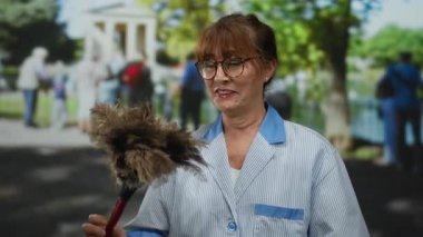 Açık hava parkında toz bezi ile gülümseyen kıdemli İspanyol kadın, sakin ve profesyonel bir temizlik ortamı öneriyor..