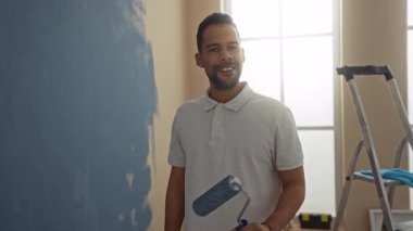 Genç, İspanyol asıllı bir adam, evin içinde yeni bir oturma odasında bir silindirle duvar boyuyor. Gülümseyerek ve çalışarak gündelik ve çekici bir ortam yaratıyor..