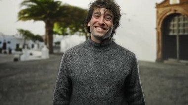 Şehir merkezinde gülümseyen genç bir adam açık havada arka planda ağaçlar olan gri bir kazak giyiyor..