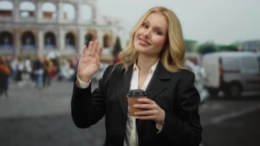 Roma, İtalya 'da kolezyumun önünde elinde kahve fincanıyla duran, hoş bir ifadeyle el sallayan ve siyah takım elbise giyen genç bir kadın, arka planda turizm manzarasının olduğunu gösteriyor..