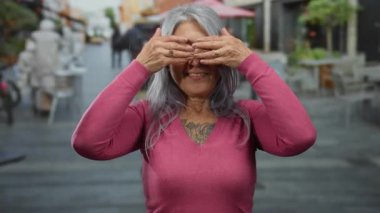 Gri saçlı kadın neşeli bir şekilde gözlerini örtüyor ve bir sokak ortamında gülümserken açık havada neşeli duygular sergiliyor..
