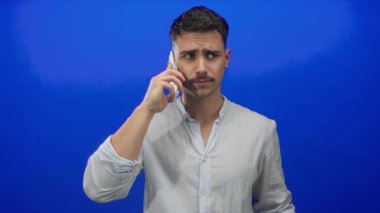 Genç İspanyol asıllı bir adam, mavi arka planda akıllı telefondan konuşuyor, çılgın jestler yapıyor, rahat bir gömlek giyiyor, hayal kırıklığını ifade ediyor, yüz ifadeleriyle duygularını iletiyor..
