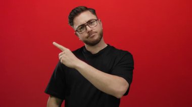 Sakallı, gözlüklü ve siyah tişörtlü genç adam izole edilmiş kırmızı duvar arka planına karşı düşünceli bir ifadeyle yukarıya bakıyor..