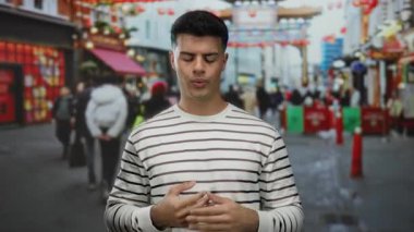 Genç adam hareketli bir şehir caddesinde gözleri kapalı, çizgili bir tişört giymiş, bulanık renkli şehir çevresi ve geçen yayalarla çevrili duruyor..